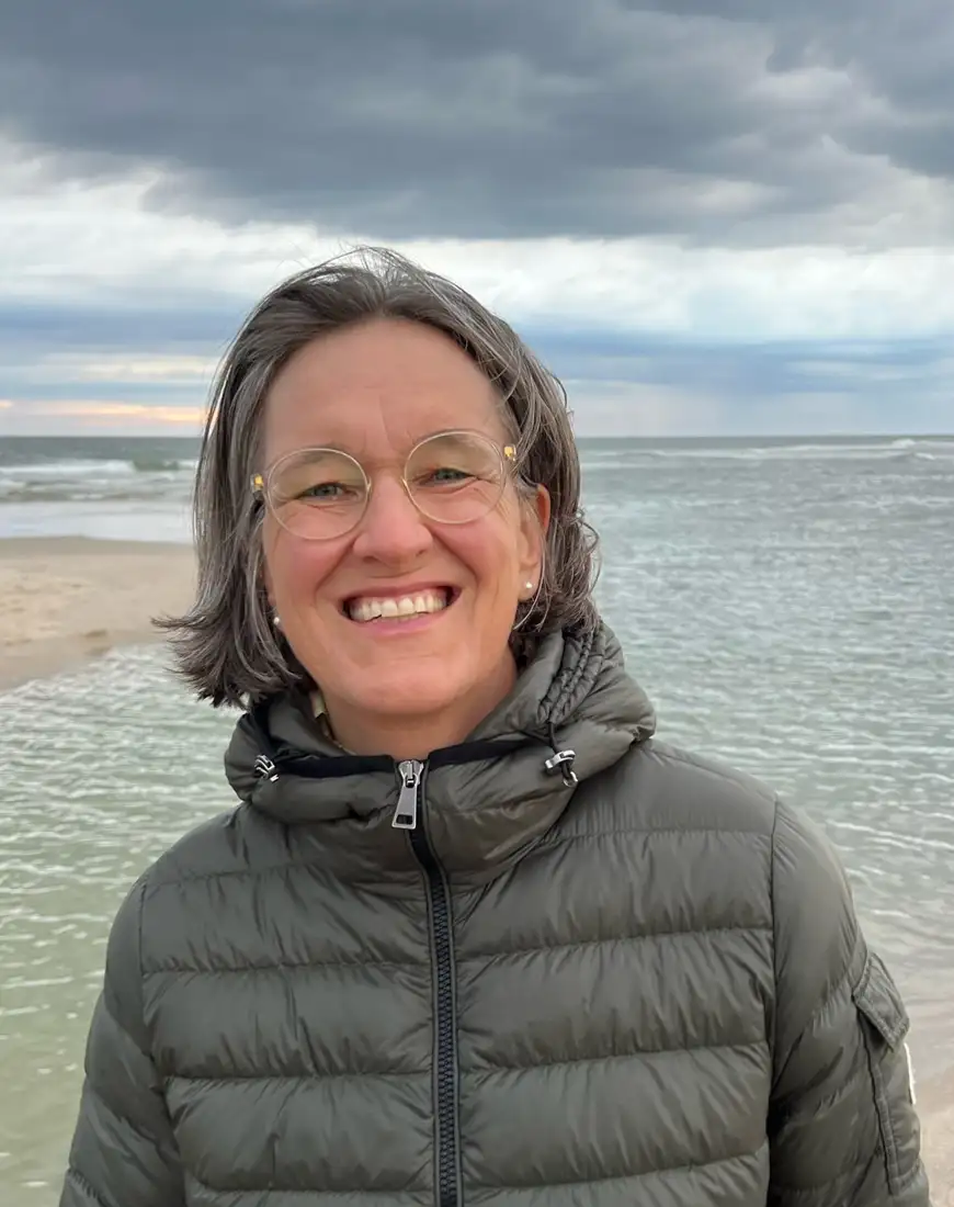 Im Hintergrund: Ein weites Meer mit Strand und bewölktem Himmel. Im Vordergrund: Eine fröhlich aussehende Frau mit einem sympathischen Lächeln.